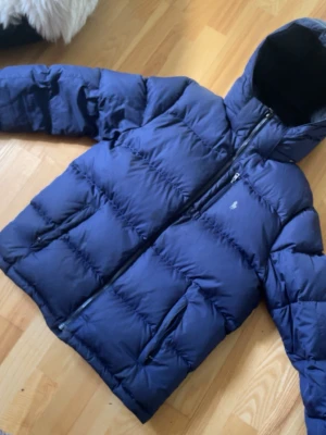 Blå pufferjacka från Polo Ralph Lauren - VÄLDIGT SÄLLSYNT Snygg blå pufferjacka från Polo Ralph Lauren med huva och dragkedja framtill. Jackan har två fickor med dragkedja och diskret logga på bröstet. Perfekt för kalla vinterdagar tack vare den tjocka vadderingen och sportiga looken.