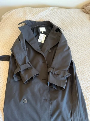 Svart trenchcoat från H&M - Stilren svart trenchcoat från H&M med klassisk dubbelknäppning, bälte i midjan och justerbara ärmslut. Jackan har bred krage och är tillverkad i ett slätt tyg som ger en snygg siluett. Perfekt för dig som gillar minimalistisk och tidlös stil. HELT NY (OANVÄND) PRISLAPP KVAR