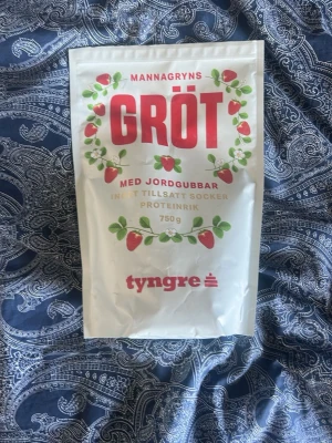 Protein gröt från tyngre - En vit påse med proteinrik mannagrynsgröt från Tyngre, smaksatt med jordgubbar och utan tillsatt socker. Förpackningen rymmer 750 gram och har röda och gröna detaljer samt jordgubbsillustrationer. Tagit en 1 skopa från paketet så typ 95% kvar