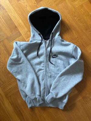 Vintage Grå zip hoodie från Nike - Snygg grå zip hoodie från Nike med svart logga på bröstet. Hoodien har dragkedja framtill, justerbar huva med svarta snören och ribbade muddar vid ärmar och midja. Insidan av huvan är svart vilket ger en schysst kontrast. Perfekt för en avslappnad streetwear-look. Fint skick 