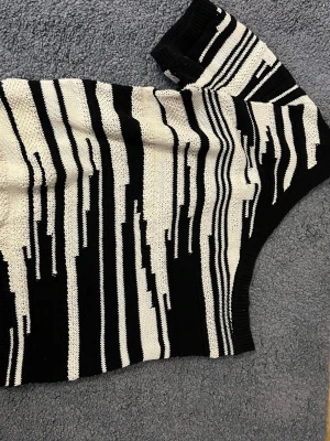 Svartvit stickad oneshoulder topp - Trendig stickad topp i svart och vitt med grafiskt randigt mönster. Modellen är oneshoulder med kort ärm.