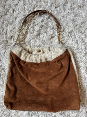 Brun handväska med fuskpäls och kedja - Säljer en snygg handväska i brun mockaimitation med mjuk vit fuskpäls upptill. Väskan har axelrem i brunt och guldfärgad kedja, samt tryckknapp för stängning. Perfekt för dig som gillar en mysig och trendig look.