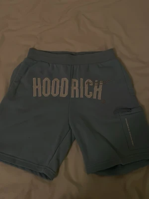 Blåa shorts från Hoodrich - Snygga blåa shorts från Hoodrich med stort tryck framtill och texten 'FROM NOTHING TO SOMETHING' på benet. Shortsen har en ficka med dragkedja på sidan och elastisk midja för skön passform. Perfekta för chill dagar eller streetwear-stil.