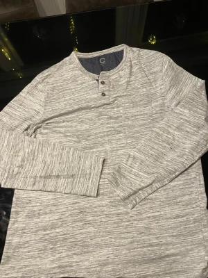 Ljusgrå långärmad henleytröja - Snygg ljusgrå långärmad henleytröja med melerad effekt och två knappar vid halsen. Tröjan har rund halsringning och är tillverkad i mjukt, stretchigt material. Perfekt för en avslappnad och stilren look.