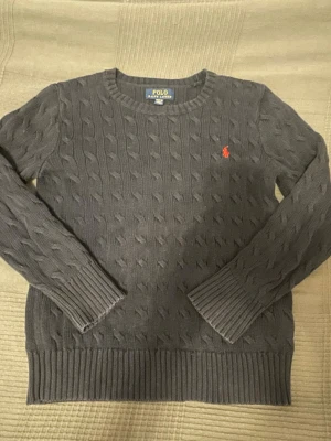 Mörkgrå kabelstickad tröja Polo Ralph Lauren - Kabelstickad mörkgrå tröja från Polo Ralph Lauren med rund halsringning och klassisk röd logga broderad på bröstet. Tröjan har långa ärmar och breda ribbade muddar vid ärmslut och nederkant. Perfekt för lager på lager och snygg till jeans.