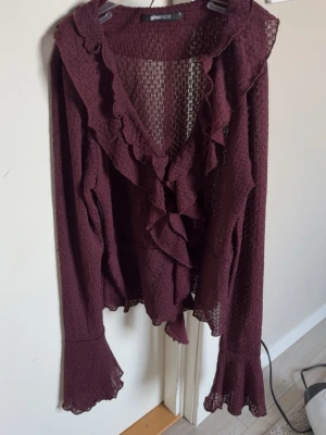 Vinröd volangblus från Gina Tricot - Vinröd blus med volangdetaljer och djup v-ringning från Gina Tricot. Blusen är i ett tunt, transparent material med struktur och har långa ärmar med utsvängda ärmslut. Perfekt för dig som gillar romantisk och trendig stil.