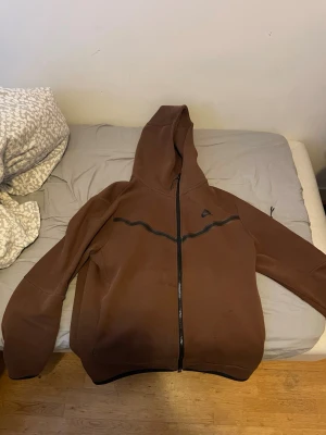 Brun hoodie med dragkedja från Nike  - Säljer en brun hoodie från Nike  med hel dragkedja framtill och svart detalj över bröstet. Hoodien har en stor huva och två fickor på sidorna. Perfekt för dig som gillar sportig och avslappnad stil. En liten grej saknas från dragkedjan!