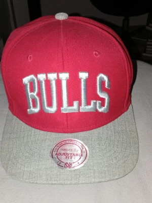 Röd Bulls keps från Mitchell & Ness - Snygg röd keps med grått flatbrätte från Mitchell & Ness. Framtill står det 'BULLS' i stora vita bokstäver och på sidan finns NBA-loggan broderad. Justerbar passform och klassisk snapback-stil. Perfekt för dig som gillar streetwear och basket.