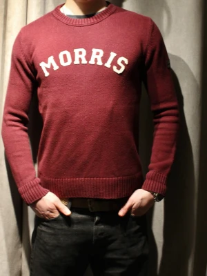 Vinröd stickad tröja från Morris - Vinröd stickad tröja från Morris med vit broderad logga på bröstet. Tröjan har rund halsringning, ribbade muddar och nederkant samt lång ärm. Perfekt för dig som gillar klassisk och stilren design.