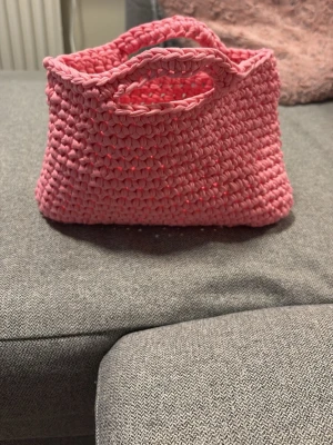 Hemmagjord virkad väska - En hemmagjord virkad väska med handtag jag själv har virkat, gjord av tjockt rosa garn. Väskan har en rektangulär form med integrerade handtag och en mjuk, chunky struktur. Perfekt för dig som gillar färgglada accessoarer och handgjorda detaljer. Passar perfekt till sommarn som en liten strandväska eller till andra tillfällen. Väskan är 23 centimeter bred, 17 centimeter hög och väskan är inte så tjock men materialet är rörligt så den anpassar sig.