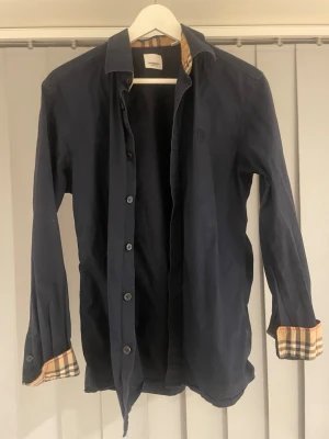 Mörkblå skjorta från Burberry - Stilren mörkblå skjorta från Burberry med klassiska rutiga detaljer på insidan av kragen och ärmsluten. Skjortan har knäppning framtill och lång ärm. Perfekt för dig som gillar exklusiva och tidlösa plagg.