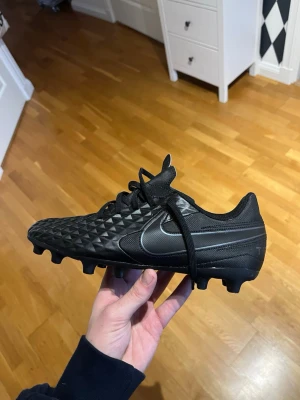 Nike Tiempo Legend 8 Club svarta fotbollsskor - Snygga svarta Nike Tiempo fotbollsskor med klassisk Swoosh-logga i silver. Skorna har en mönstrad ovandel i syntetmaterial för extra grepp och stabilitet samt fasta dobbar för spel på gräs. Perfekta för dig som vill ha stilrena och funktionella skor på planen. Knappt använda!