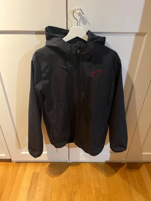 Svart vindjacka från doubles  - Svart vindjacka med huva och dragkedja framtill. På bröstet finns ett rött hjärta som detalj. Jackan har justerbara snören vid huvan och är tillverkad i ett lätt, vindtätt material. Perfekt för blåsiga dagar och enkel att matcha med olika outfits.