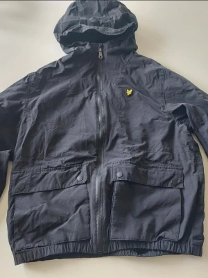 Svart vindjacka från Lyle & Scott - Snygg svart vindjacka från Lyle & Scott med huva och dragkedja framtill. Jackan har två stora fickor med knappar och det klassiska gula logomärket på bröstet. Tillverkad i lätt och slitstarkt material med meshfoder för extra komfort.
