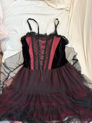 Svart och vinröd korsettklänning  - Säljer en snygg svart och vinröd korsettklänning med spetsdetaljer och snörning framtill. Klänningen har tunna axelband, volangig kjol i flera lager tyll och sammetspaneler. Den har dock en deffekt som syns på sista bilden men det går bort om man rengör försiktigt. 