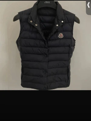 Svart dunväst från Moncler - Stilren svart dunväst från Moncler med klassisk krage och tryckknappar framtill. Västen har ett diskret Moncler-märke på bröstet och är tillverkad i glansigt material med quiltad design. Perfekt för lager-på-lager och snygg till streetwear.