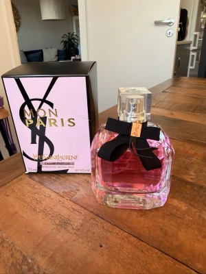Yves Saint Laurent Mon Paris parfym - Yves Saint Laurent Mon Paris Eau de Parfum i en elegant, fyrkantig glasflaska med rosa doftvätska. Flaskan har en dekorativ svart rosett med gulddetalj vid halsen och kommer med originalkartong i rosa och svart.Karongen öppnad och endast provdoftat,tyvärr ej min doft