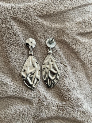Silvriga örhängen i metall - Säljer ett par coola silvriga örhängen med unik, skulpterad och organisk form. Örhängena är gjorda i metall och har en matt finish med textur som ger en modern och edgy känsla. Perfekta för dig som gillar att sticka ut med dina accessoarer.