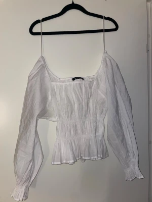 Vit blus med puffärm och smock - Snygg vit blus med smockad midja och långa puffärmar. Toppen har tunna axelband och volangkant nertill för en romantisk vibe. Perfekt att styla med jeans eller kjol för en trendig look.