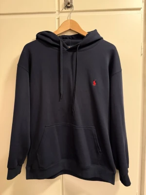 Mörkblå hoodie från Polo Ralph Lauren - Snygg mörkblå hoodie från Polo Ralph Lauren med klassisk röd logga broderad på bröstet. Hoodien har huva med dragsko, stor magficka och ribbade muddar vid ärmar och nederkant. Tillverkad i mjukt bomullsmaterial som känns skönt mot huden. Storleken är L men passar som M.