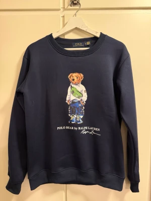Marinblå tröja Polo Ralph Lauren - Marinblå långärmad tröja från Polo Ralph Lauren med ikoniskt Polo Bear-tryck framtill. Tröjan har rund halsringning och ribbade muddar vid ärmslut och nederkant. Perfekt för dig som gillar streetstyle och klassiska märken.