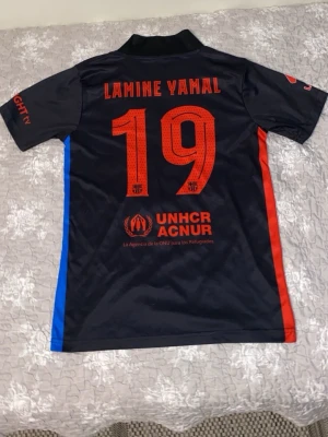FC Barcelona matchtröja Lamine Yamal - Snygg svart FC Barcelona matchtröja med Lamine Yamal och nummer 19 på ryggen. Tröjan har röda och blå detaljer på sidorna, klubbmärke på bröstet och sponsorer som Spotify och UNHCR. Tillverkad i lätt och ventilerande polyester, perfekt för fotbollsträning.