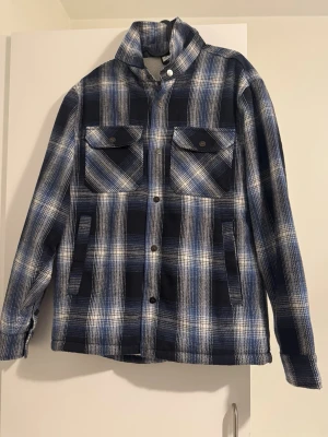 Blåvit rutig overshirt med knappar - Overshirt i blått och vitt rutigt mönster med knappar framtill och två bröstfickor med lock. Jackan har en rak passform och är perfekt för lager-på-lager. Materialet känns mjukt och värmande, troligen bomull eller ullblandning.