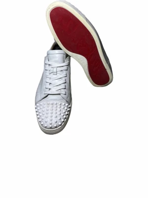 Vita Louboutin sneakers med nitar - Snygga vita sneakers från Christian Louboutin i läder med klassiska röda sulor och coola nitar på tån. Skorna har lågt skaft, vita skosnören och en clean design som sticker ut med sin edgy detalj framtill.