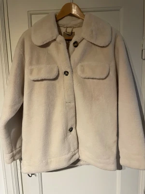Beige pälsjacka med stora fickor - Mysig beige pälsjacka i fuskpäls med bred krage och stora bröstfickor. Jackan har knäppning med knappar framtill och en rak, avslappnad passform. Perfekt för dig som gillar en enkel men trendig stil. Materialet är mjukt och fluffigt.