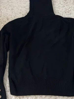 Svart stickad tröja från H&M - Snygg svart stickad tröja från H&M med hög krage och ribbade muddar. Tröjan har långa ärmar och dekorativa knappar vid ärmslutet. En knapp saknas på ene ärmet