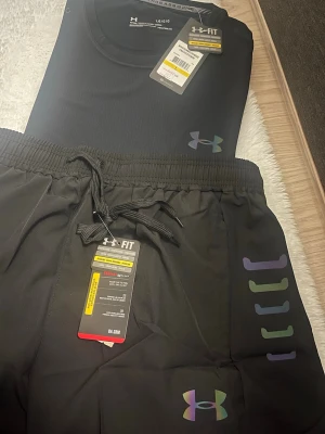 Svart träningsset från Under Armour - Snyggt svart träningsset från Under Armour med t-shirt och shorts. Båda plaggen har regnbågsfärgade loggor och textdetaljer. Materialet är lätt och andas, perfekt för träning eller sport. Elastisk midja med dragsko på shortsen för skön passform.