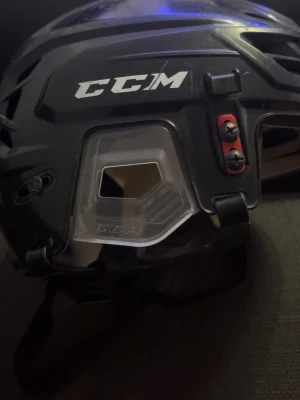 Svart hockeyhjälm från CCM - Säljer en svart hockeyhjälm från CCM med justerbar passform och transparenta detaljer på sidorna. Hjälmen har ventilationshål, hakrem och tydlig logga på sidan. Perfekt för dig som spelar ishockey och vill ha bra skydd.