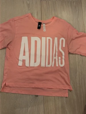 Rosa Adidas t-shirt med stor logga - Snygg rosa t-shirt från Adidas med stor vit logga framtill. T-shirten har rund halsringning och korta ärmar. Tillverkad i mjuk bomull som känns skön mot huden. Perfekt för en avslappnad och sportig stil.