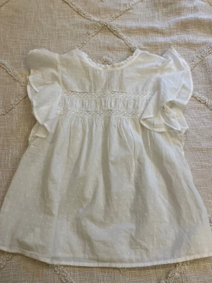 Vit blus med volangärmar och broderi - Söt vit blus med diskret prickmönster och volangiga kortärmar. Blusen har fina broderidetaljer framtill och en liten knapp i nacken. Tillverkad i ett lätt och luftigt bomullstyg, perfekt för en fräsch och somrig look. Använd bara en gång💞