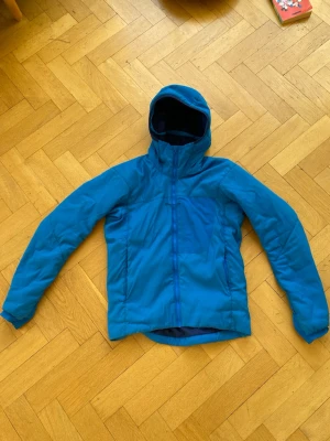 Arc'teryx proton - Blå pufferjacka från Arc'teryx med huva och dragkedja framtill. Jackan har en sportig look och är tillverkad i slitstark polyester, perfekt för kalla dagar. Fodret är mörkblått och jackan har en clean design utan onödiga detaljer.