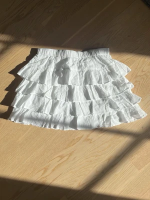 Vit volangkjol från Kappahl - Säljer en superfin vit kjol från Kappahl med flera lager volanger. Kjolen har resår i midjan och är gjord i ett lätt och luftigt material, perfekt för varma dagar. Enkel att matcha med olika toppar för en trendig look.