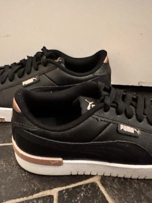 Svarta Puma sneakers med kopparfärg - Snygga svarta sneakers från Puma med detaljer i kopparfärg på hälen och logga på sidan. Skorna har en vit sula och är tillverkade i ett slitstarkt material med klassisk låg profil och snörning. Perfekta för dig som vill ha en stilren och sportig look. Använda en gång