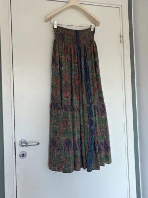 Grön paisleymönstrad långkjol - Säljer en flowy grön långkjol med paisleyinspirerat mönster i rött, blått och beige. Kjolen har resår i midjan och ett brett fall, perfekt för dig som gillar bohemiska vibes. Vet inte märket men den är köpt i Italien 