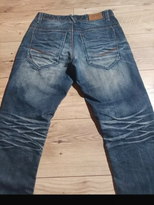 Blå jeans med slitningar och kontrastsömmar - Säljer ett par blå jeans med snygga slitningar och markerade kontrastsömmar. Jeansen har klassisk femficksmodell och coola detaljer på bakfickorna. Perfekta för en avslappnad och trendig stil.