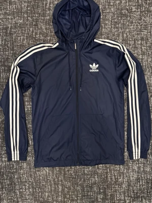 Marinblå Adidas vindjacka med huva - Snygg marinblå vindjacka från Adidas med klassiska vita ränder längs ärmarna och vit logga på bröstet. Jackan har huva, dragkedja framtill och två fickor. Perfekt för en sportig och avslappnad stil.