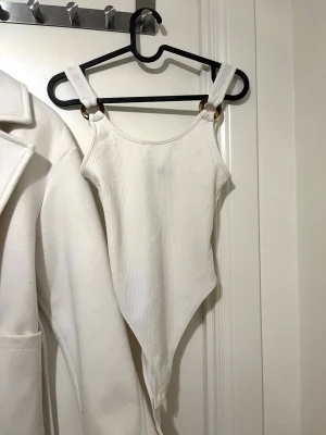 Vit ribbad body med breda axelband - Snygg vit ribbad body med breda axelband och guldfärgade ringdetaljer framtill. Bodyn har en spetsig nederdel och är tillverkad i stretchigt, mjukt material. Perfekt att styla med jeans eller kjol för en trendig look.