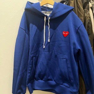 Blå hoodie Comme des Garçons Play - Säljer en blå hoodie från Comme des Garçons Play med den ikoniska röda hjärt-loggan på bröstet. Tröjan har huva med vit snörning och känguruficka framtill. Perfekt för dig som gillar streetwear och vill sticka ut lite extra. Står storlek S, men sitter som Xs.