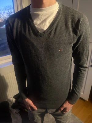 Mörkgrå V-ringad tröja Tommy Hilfiger - Snygg mörkgrå V-ringad tröja från Tommy Hilfiger med diskret logga på bröstet. Tröjan har lång ärm och är tillverkad i ett tunt, mjukt material, säljer nu den här för att den är för liten|modellen är i 177cm lång| nypris 1249