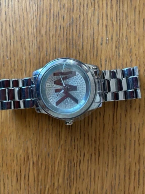 Silverfärgad Michael Kors klocka - Snygg klocka från Michael Kors i silverfärgat metallarmband. Urtavlan har stora MK-bokstäver och är dekorerad med glittriga stenar för en extra touch. Klassisk rund form och robust länkarmband gör den till ett stilrent val.