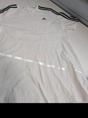 Vit Adidas t-shirt med svarta ränder - Klassisk vit t-shirt från Adidas med svarta ränder på axlarna och det ikoniska Adidas-loggan broderad på bröstet. T-shirten har rund hals och är tillverkad i mjuk bomull, perfekt för en sportig och avslappnad stil.