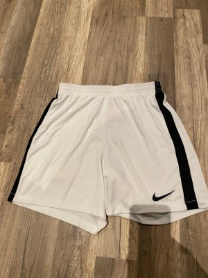 Vita Nike shorts med svarta detaljer - Snygga vita shorts från Nike med svarta ränder längs sidorna och klassisk Nike-logga på vänster ben. Shortsen har elastisk midja och är tillverkade i ett lätt och svalt syntetmaterial, perfekta för sport och träning.