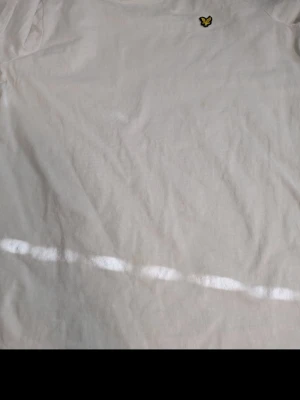 Vit t-shirt från Lyle & Scott - Säljer en klassisk vit t-shirt från Lyle & Scott med den ikoniska gula loggan på bröstet. T-shirten är kortärmad och gjord i mjuk bomull, perfekt för en clean och enkel stil.