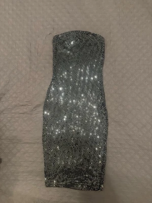 Glittrig svart axelbandslös klänning - Säljer en svart, glittrig axelbandslös kortklänning med tajt passform. Klänningen är täckt av små silverfärgade paljetter som ger en riktigt festlig look. Perfekt för dig som vill sticka ut och glänsa på nästa event!
