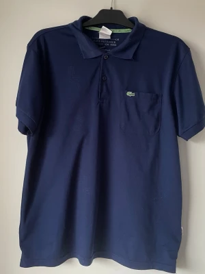 Marinblå pikétröja från Lacoste storlek 7 - Klassisk marinblå pikétröja från Lacoste med korta ärmar, krage och knappar framtill. Tröjan har en bröstficka med den ikoniska gröna krokodilloggan. Perfekt till en avslappnad men ändå stilren look. Storlek 7