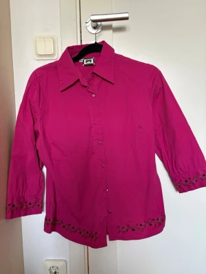 Ceriserosa blus  - Snygg ceriserosa blus från JOY. Blusen har fina svarta blombroderier längs ärmslut och nederkant som ger en fin touch. 